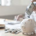 White piggy bank with a smiling face, hand holding cash, and scattered coins for HVAC financial planning.