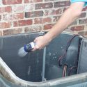 Hand holding a handheld sprayer near a container with wiring against a brick wall in HVAC maintenance setting.